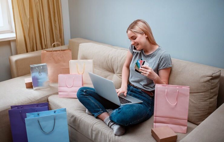 Foto joven comprador sonriente con laptop y tarjetas de crédito está ordenando por teléfono inteligente en una tienda en línea mientras está sentado en un sofá con bolsas de compras
